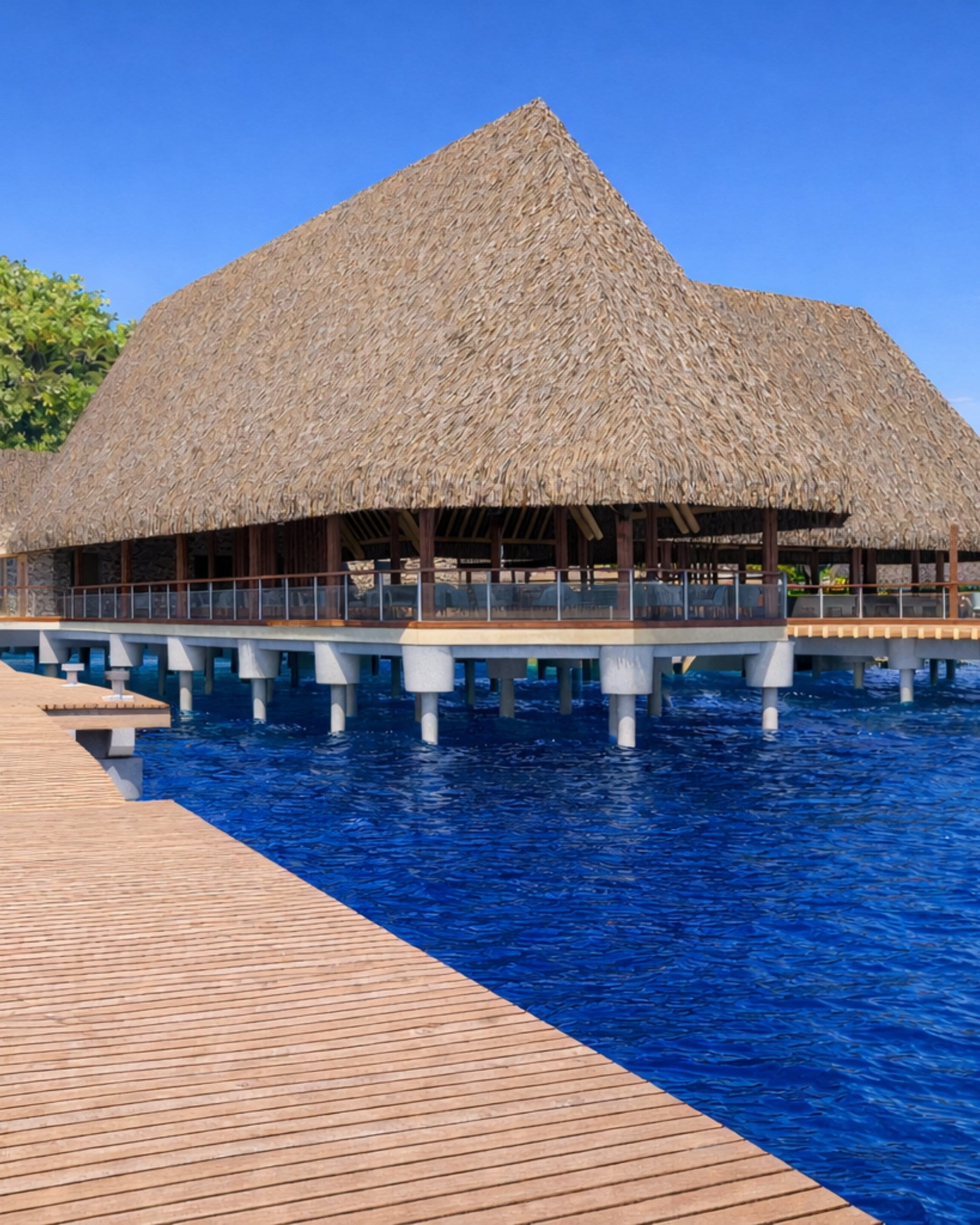 Grand pavillon de restaurant sur pilotis avec toit de chaume au-dessus du lagon, passerelle en bois