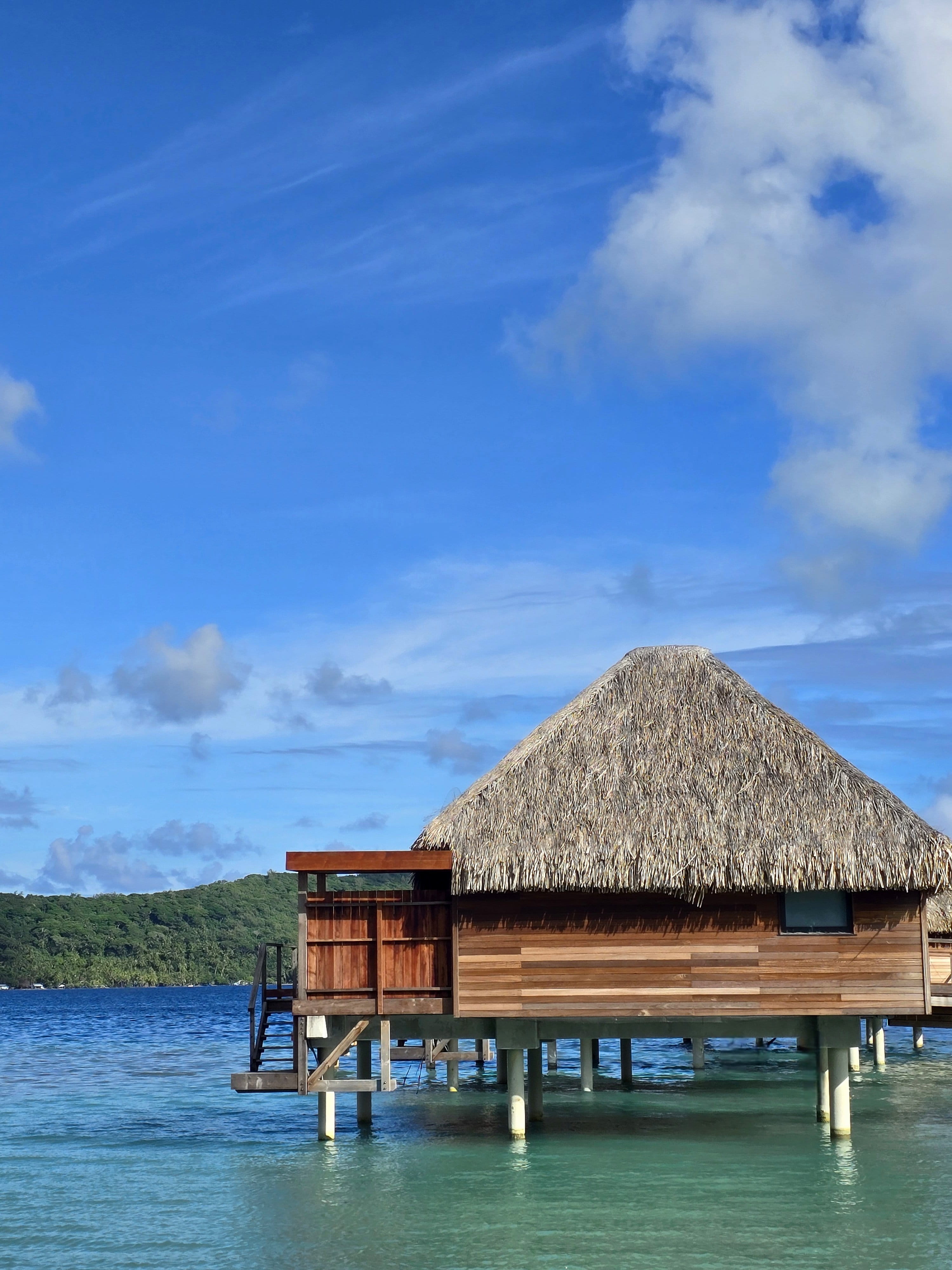 Bungalow sur pilotis au toit de chaume au-dessus d'un lagon turquoise, île verdoyante et ciel bleu.